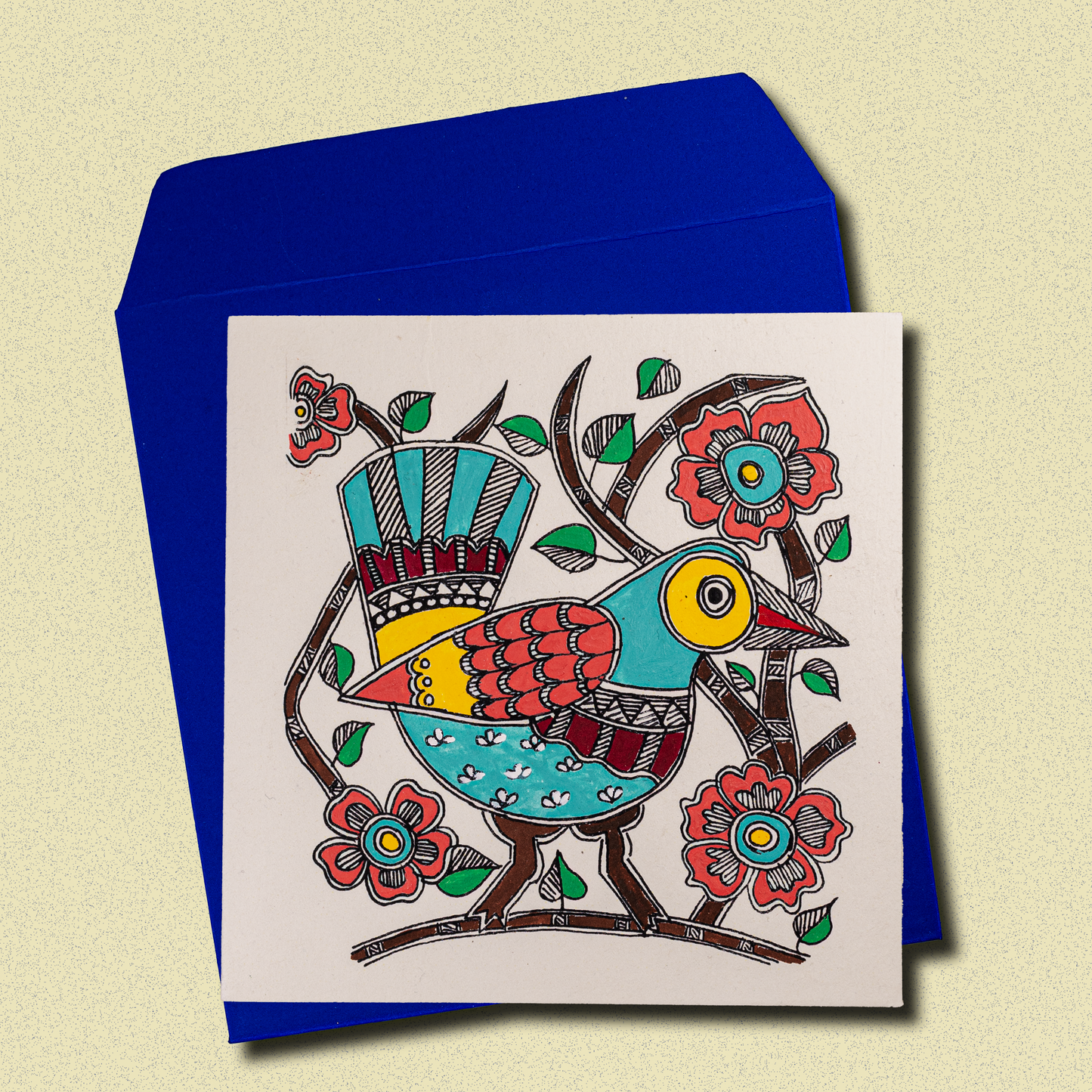 Colorful hand-painted Madhubani-style greeting card featuring a vibrant folk art bird with decorative patterns, surrounded by stylized flowers and branches, displayed with a blue envelope on a beige background