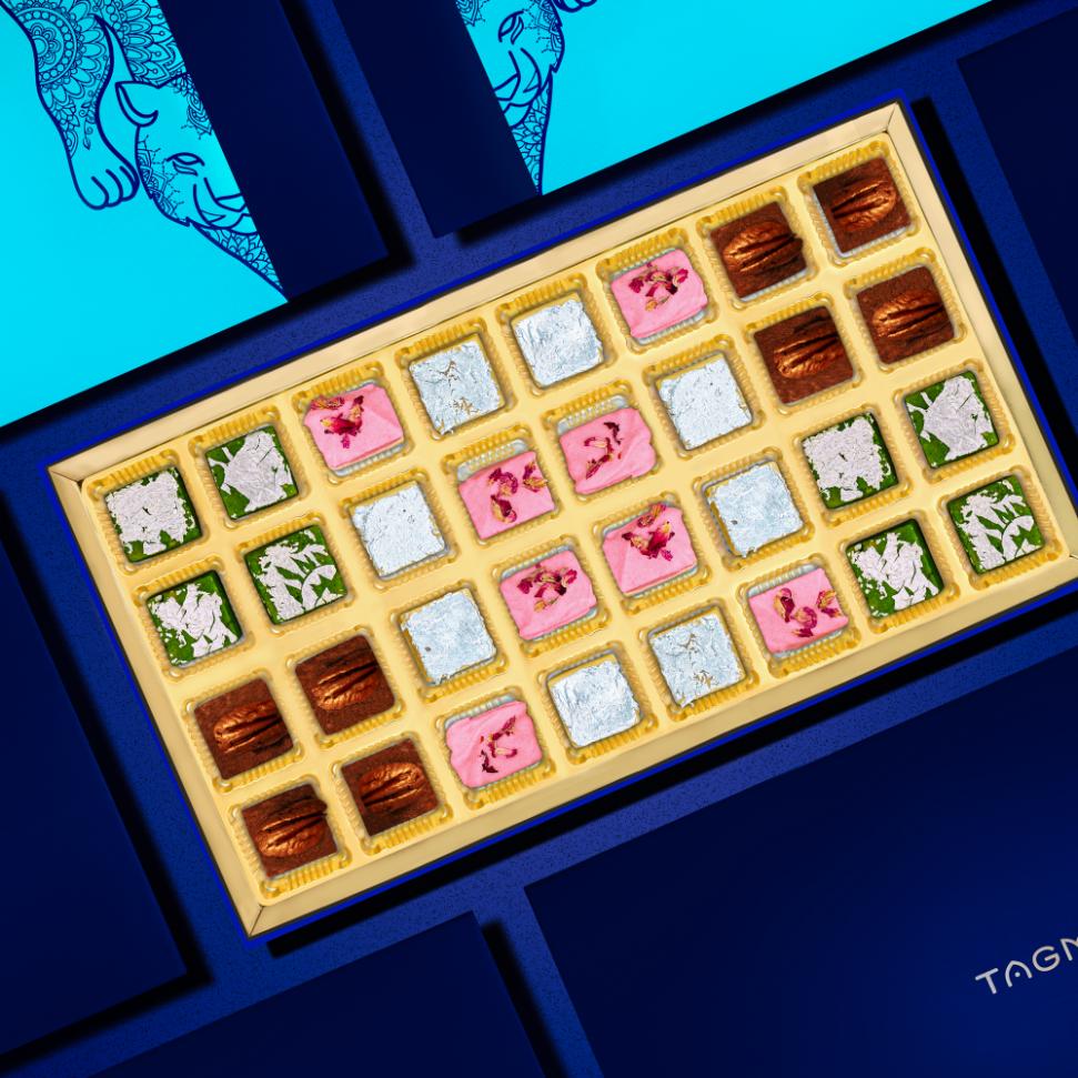 
                      
                        Colorful assortment of Indian burfi sweets in a luxurious box, featuring various flavors like pistachio, rose, and pecan, displayed on a vibrant blue background with intricate henna-inspired designs.
                      
                    