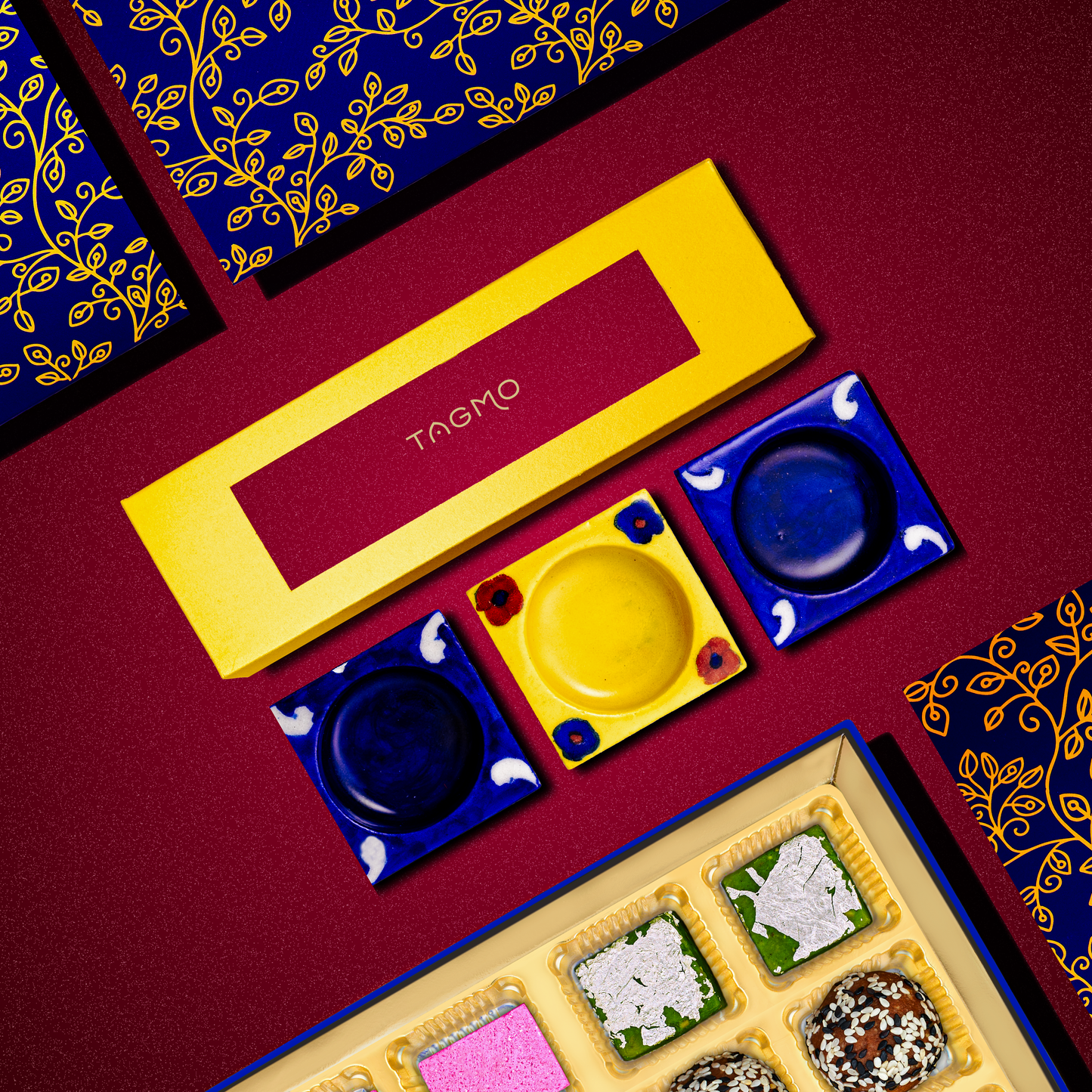 Hand-painted blue and yellow porcelain diyas in a set of three, displayed with decorative packaging and traditional Indian sweets on a burgundy background with gold-patterned fabric, showcasing artisanal craftsmanship from Jaipur