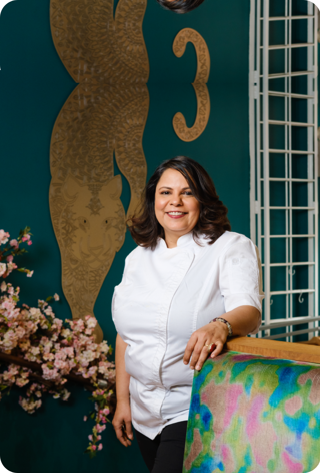 Woman in a white coat standing next to a colorful painting with decorative elements in the background.