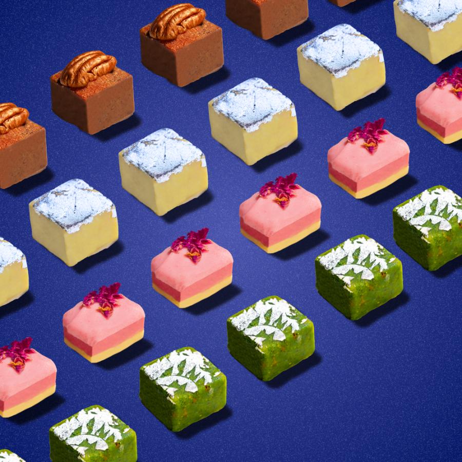Assorted Indian burfi sweets arranged in diagonal rows on a blue background, showcasing various flavors including pecan, pistachio, rose, and coconut. The colorful square-shaped confections display different toppings and textures, highlighting the diversity of the Chef's Burfi Collection.