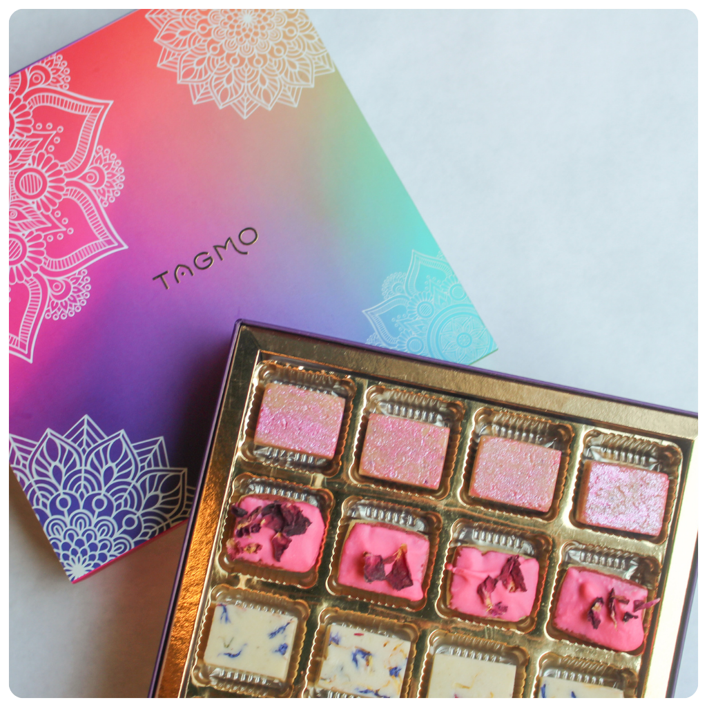 A colorful assortment of traditional Indian sweets, known as mithai, are displayed in a decorative box. The box features a vibrant, mandala-inspired design in various shades of pink, purple, and blue. The mithai appear to be a variety of flavors, including rose, coconut, lavender, and thandai.